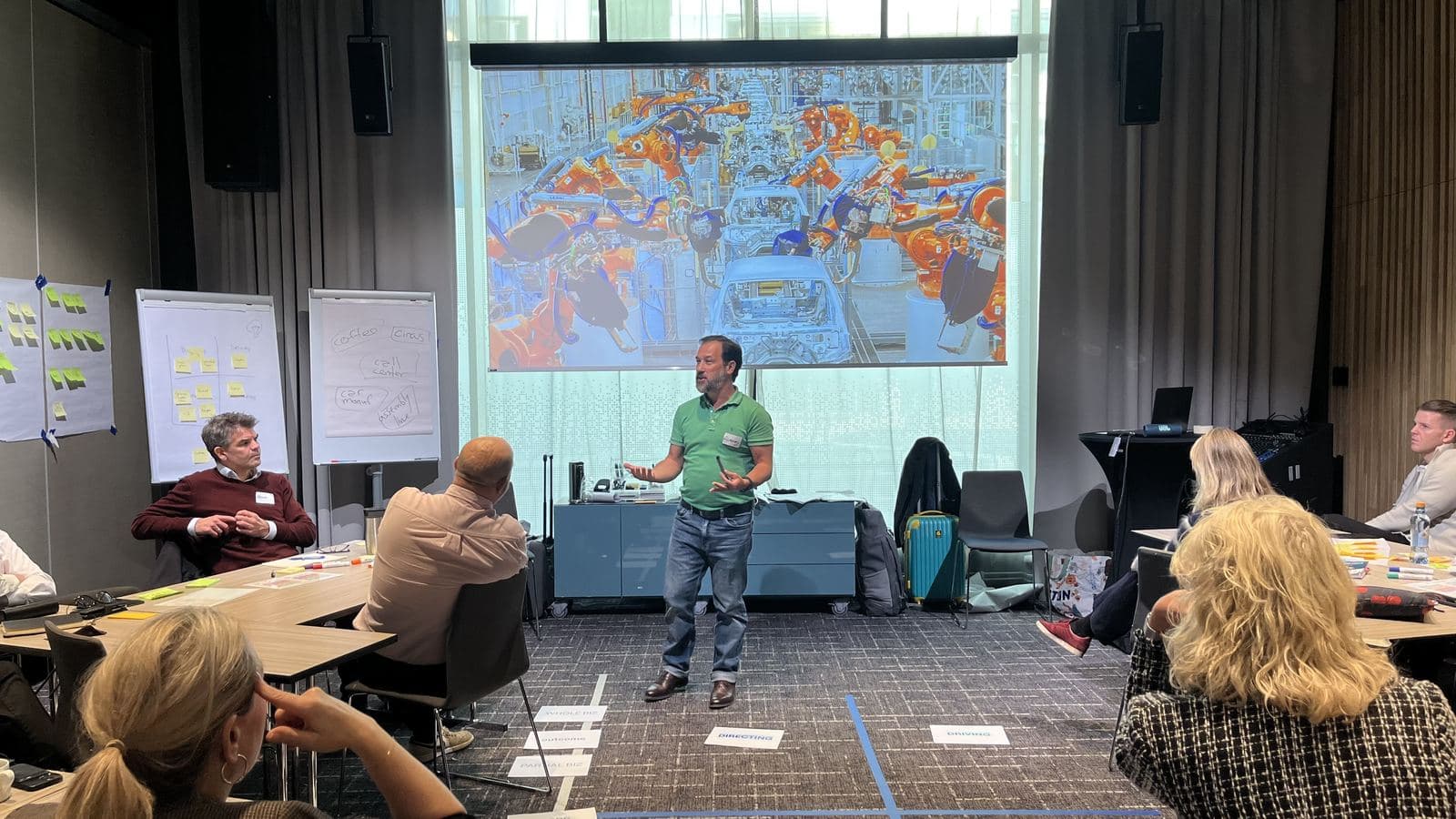 Alexey Krivitsky standing mid-session in front of participants seated at tables, flipcharts covered in sticky notes along the left wall, and a car-factory robot assembly line on the projection screen behind him.